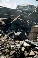 Charred and collapsed wooden beams and debris are scattered across a pile, with a partially damaged roof visible in the background. The scene suggests destruction, likely from a fire.