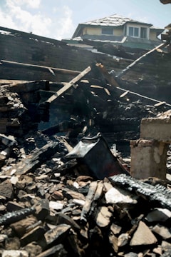Charred and collapsed wooden beams and debris are scattered across a pile, with a partially damaged roof visible in the background. The scene suggests destruction, likely from a fire.