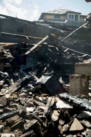 Charred and collapsed wooden beams and debris are scattered across a pile, with a partially damaged roof visible in the background. The scene suggests destruction, likely from a fire.