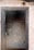 A doorway heavily charred by fire leads into an interior space with smoke-stained tiles and water fixtures. The framing of the door appears severely burnt, and black soot marks are visible around the entry. A wire hangs loosely from the doorframe, indicating possible electrical damage.