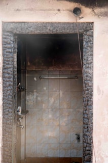 A doorway heavily charred by fire leads into an interior space with smoke-stained tiles and water fixtures. The framing of the door appears severely burnt, and black soot marks are visible around the entry. A wire hangs loosely from the doorframe, indicating possible electrical damage.