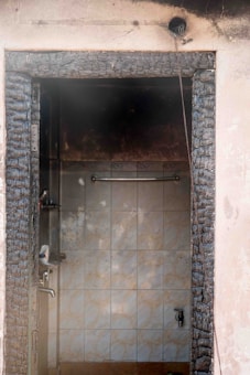 A doorway heavily charred by fire leads into an interior space with smoke-stained tiles and water fixtures. The framing of the door appears severely burnt, and black soot marks are visible around the entry. A wire hangs loosely from the doorframe, indicating possible electrical damage.