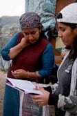 Two women are engaged in a focused discussion, holding papers in their hands. One woman wears a colorful headscarf and a maroon vest over a blue top, while the other wears a light-colored cap and a dark hoodie. They appear to be outdoors with a natural landscape in the background.
