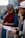 Two women are engaged in a focused discussion, holding papers in their hands. One woman wears a colorful headscarf and a maroon vest over a blue top, while the other wears a light-colored cap and a dark hoodie. They appear to be outdoors with a natural landscape in the background.