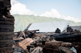 A collapsed structure with rubble and debris is present in the foreground, with a backdrop of green hills and a bright blue sky. The remains of the building include charred wooden beams, scattered stones, and a partial roof covered by a white tarp, suggesting previous damage by fire or a natural disaster.