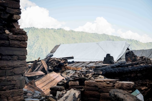 A collapsed structure with rubble and debris is present in the foreground, with a backdrop of green hills and a bright blue sky. The remains of the building include charred wooden beams, scattered stones, and a partial roof covered by a white tarp, suggesting previous damage by fire or a natural disaster.