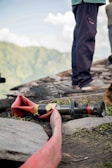 A coiled red fire hose is laid out on the ground near a rocky surface, accompanied by a nozzle attachment. The scene includes sections of a person standing nearby, wearing dark pants and shoes. A blurred, lush green landscape is visible in the background under a partly cloudy sky.