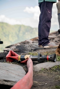 A coiled red fire hose is laid out on the ground near a rocky surface, accompanied by a nozzle attachment. The scene includes sections of a person standing nearby, wearing dark pants and shoes. A blurred, lush green landscape is visible in the background under a partly cloudy sky.