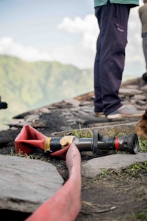 A coiled red fire hose is laid out on the ground near a rocky surface, accompanied by a nozzle attachment. The scene includes sections of a person standing nearby, wearing dark pants and shoes. A blurred, lush green landscape is visible in the background under a partly cloudy sky.