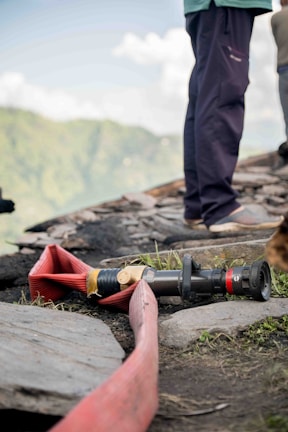A coiled red fire hose is laid out on the ground near a rocky surface, accompanied by a nozzle attachment. The scene includes sections of a person standing nearby, wearing dark pants and shoes. A blurred, lush green landscape is visible in the background under a partly cloudy sky.