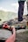 A coiled red fire hose is laid out on the ground near a rocky surface, accompanied by a nozzle attachment. The scene includes sections of a person standing nearby, wearing dark pants and shoes. A blurred, lush green landscape is visible in the background under a partly cloudy sky.