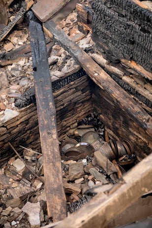 A collection of metal objects and debris are found among charred wooden beams and rubble. The scene appears to depict remnants of a burned or collapsed structure, with visible blackened wood and scattered stones.