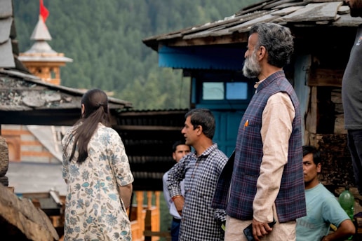 A group of people stand outdoors near rustic wooden structures with a backdrop of green trees and a distant building with a red flag on top. The individuals are engaged in conversation and appear to be in a rural setting.