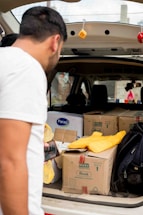 A person is leaning into the back of an open vehicle, which is filled with cardboard boxes, some labeled with brand names. A yellow fabric item and a black backpack are also visible, along with hanging decorative dice from the vehicle's roof. The person is holding a smartphone.