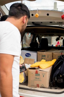A person is leaning into the back of an open vehicle, which is filled with cardboard boxes, some labeled with brand names. A yellow fabric item and a black backpack are also visible, along with hanging decorative dice from the vehicle's roof. The person is holding a smartphone.