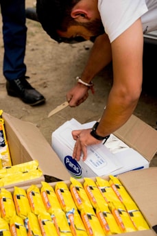 A person is using a knife to open a cardboard box labeled 'Paras'. Next to this box, there are several packets of yellow-packaged food items. The action takes place outdoors on a dirt ground.