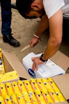 A person is using a knife to open a cardboard box labeled 'Paras'. Next to this box, there are several packets of yellow-packaged food items. The action takes place outdoors on a dirt ground.