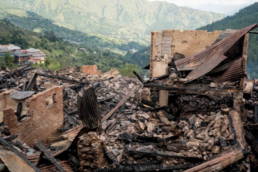 A scene of devastation showing the ruins of a building. The structure appears severely damaged, with collapsed walls and charred debris, likely indicating a fire or disaster. The background reveals a lush, green mountainous landscape with additional buildings visible in the distance.