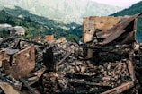Rubble from a collapsed or burnt building is spread across the foreground, characterized by broken bricks, charred wood, and partially remaining structures. In the background, a lush green mountainous landscape can be seen, providing a stark contrast to the devastation in the foreground.
