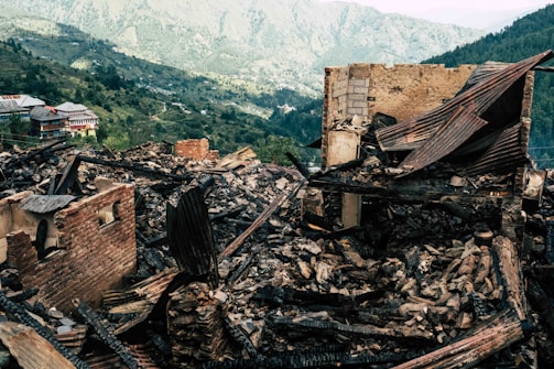 Rubble from a collapsed or burnt building is spread across the foreground, characterized by broken bricks, charred wood, and partially remaining structures. In the background, a lush green mountainous landscape can be seen, providing a stark contrast to the devastation in the foreground.
