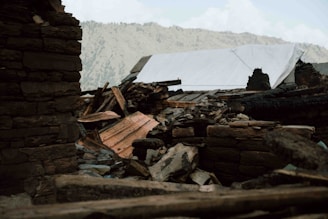 Rubble from a collapsed structure, including charred wood and large stones, sits in the foreground. A makeshift tarp covers part of the debris. Mountainous terrain is visible in the background under a cloudy sky.