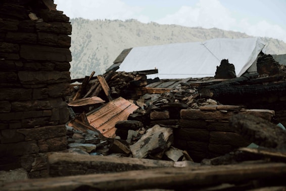 Rubble from a collapsed structure, including charred wood and large stones, sits in the foreground. A makeshift tarp covers part of the debris. Mountainous terrain is visible in the background under a cloudy sky.