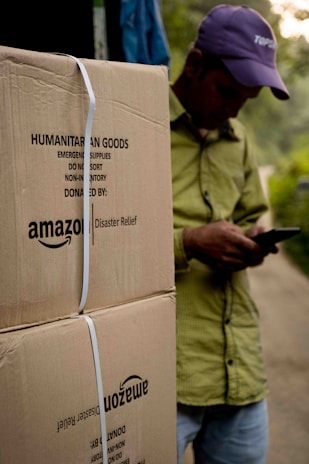 A person wearing a hat is leaning against large cardboard boxes labeled 'Amazon Disaster Relief' with humanitarian goods. The individual appears to be using a mobile phone. The boxes are strapped and suggest they are ready for transport or distribution. The setting appears to be outdoors with some greenery in the background.