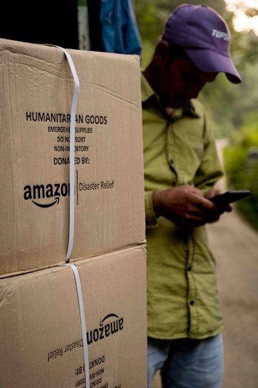 A person wearing a hat is leaning against large cardboard boxes labeled 'Amazon Disaster Relief' with humanitarian goods. The individual appears to be using a mobile phone. The boxes are strapped and suggest they are ready for transport or distribution. The setting appears to be outdoors with some greenery in the background.