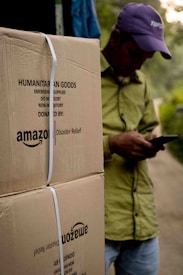 A person wearing a hat is leaning against large cardboard boxes labeled 'Amazon Disaster Relief' with humanitarian goods. The individual appears to be using a mobile phone. The boxes are strapped and suggest they are ready for transport or distribution. The setting appears to be outdoors with some greenery in the background.