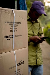 A person wearing a hat is leaning against large cardboard boxes labeled 'Amazon Disaster Relief' with humanitarian goods. The individual appears to be using a mobile phone. The boxes are strapped and suggest they are ready for transport or distribution. The setting appears to be outdoors with some greenery in the background.