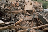 A scene of devastation with charred wooden remains and bricks scattered throughout the area. The remnants of a structure are visible with walls partially standing amidst the debris. The ground is covered in ash and burned materials, indicating a fierce fire has previously swept through.