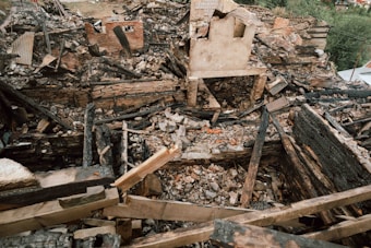 A scene of devastation with charred wooden remains and bricks scattered throughout the area. The remnants of a structure are visible with walls partially standing amidst the debris. The ground is covered in ash and burned materials, indicating a fierce fire has previously swept through.