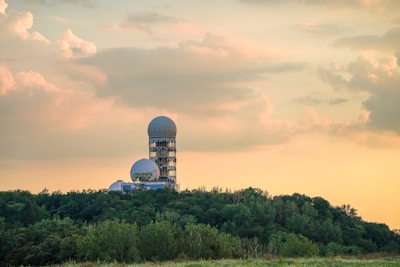 A large structure with geodesic domes rises above a dense forest, set against a sky with scattered clouds illuminated with warm, golden light. Part of the structure bears graffiti, adding an urban contrast to the natural surroundings.