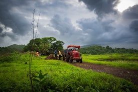 A rural landscape features a red tractor on a dirt path surrounded by lush green fields under a cloudy sky. Several people are working near the tractor, digging or handling soil. Trees and distant hills form the background, contributing to a dynamic and slightly dramatic atmosphere.