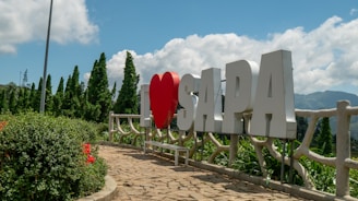 Large white letters spell out 'I ❤️ SAPA' against a backdrop of lush green trees and clear blue sky. A paved pathway leads up to the sign, bordered by vibrant flower bushes.