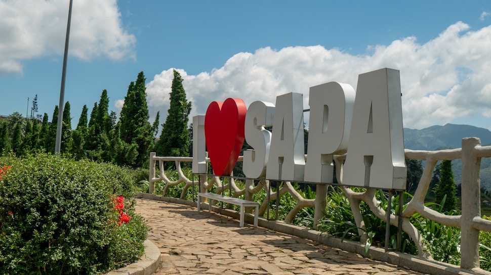 Large white letters spell out 'I ❤️ SAPA' against a backdrop of lush green trees and clear blue sky. A paved pathway leads up to the sign, bordered by vibrant flower bushes.