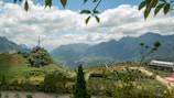 The image depicts a scenic mountain landscape with lush greenery and terraced fields. In the foreground, there are trees and plants framing the view. A telecommunications tower is situated on a small hill, and a paved path meanders through the fields. The background showcases a range of mountains under a sky filled with fluffy clouds.