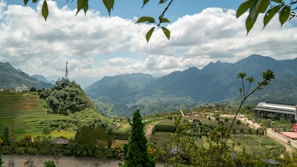 The image depicts a scenic mountain landscape with lush greenery and terraced fields. In the foreground, there are trees and plants framing the view. A telecommunications tower is situated on a small hill, and a paved path meanders through the fields. The background showcases a range of mountains under a sky filled with fluffy clouds.