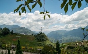 The scene depicts a lush, mountainous landscape with a prominent peak at the center, bordered by terraced fields and valleys. A notable structure or statue stands on the central peak. The foreground includes clusters of trees and small structures, possibly huts or gazebos. Leaves hang down from the top of the image, framing the view, while a bright, partly cloudy sky provides a majestic backdrop. The word 'FANSIPAN' is visible on one of the hills.