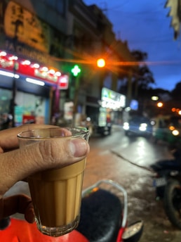 A hand holding a glass of tea is prominently displayed in the foreground, while the background shows a busy street scene at dusk with bright shop signs, streetlights, and blurred vehicles passing by.
