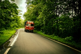 A red truck is driving down a winding road through a lush green forest. The road is wet, indicating recent rain, and is flanked by dense trees on both sides. The truck is labeled 'Goods Carrier' and appears to be traveling smoothly along the road.