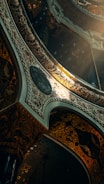 Elaborate architectural design featuring intricate patterns and ornate details, illuminated by a warm light source. The structure includes arches and decorative elements with a blend of dark and luminous tones.