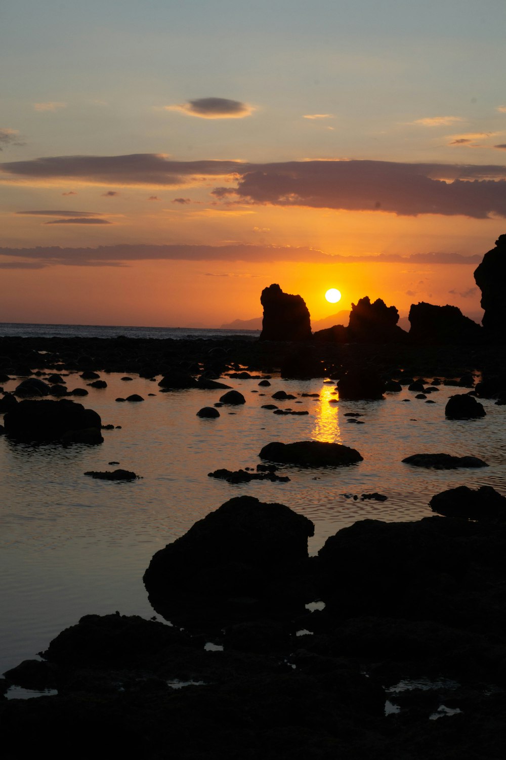 the sun is setting over the ocean with rocks in the foreground