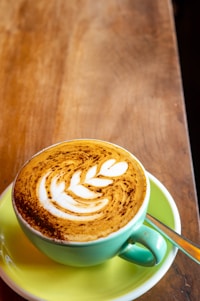 A beautifully crafted cup of coffee with latte art in the shape of a leaf sits on a wooden table. The cup is turquoise and rests on a yellow saucer, with a silver spoon placed beside it.