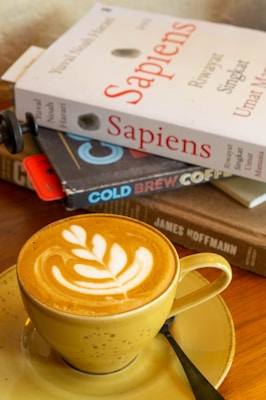 A cappuccino with latte art sits in a yellow cup and saucer. Behind it, there is a stack of books including a visible copy of 'Sapiens'. The setting suggests a cozy, leisurely atmosphere with a focus on reading and enjoying coffee.