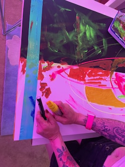 A person with tattoos on their arm is creating an abstract painting on a table. They hold a yellow marker and a black pen, working on a vibrant artwork featuring bold splashes of red, pink, green, and blue colors. Some art tools and supplies are visible nearby.