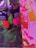 Several tubes of Liquitex Basics Acrylic paint stand on a colorful canvas. The colors include vibrant greens, pinks, and oranges. The canvas underneath features abstract, energetic brushstrokes in vivid pink, red, and orange hues, with some areas of dark green and black.
