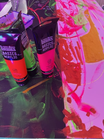 Several tubes of Liquitex Basics Acrylic paint stand on a colorful canvas. The colors include vibrant greens, pinks, and oranges. The canvas underneath features abstract, energetic brushstrokes in vivid pink, red, and orange hues, with some areas of dark green and black.