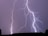Several vivid lightning bolts illuminate a dark, overcast night sky, with some trees visible in silhouette at the bottom.