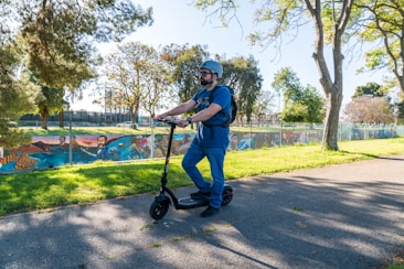 A person wearing a helmet and sunglasses is riding an electric scooter along a paved path in a park. Surrounding the path are trees and a fence with colorful graffiti. The scene is bright and sunny, with shadows cast on the ground.